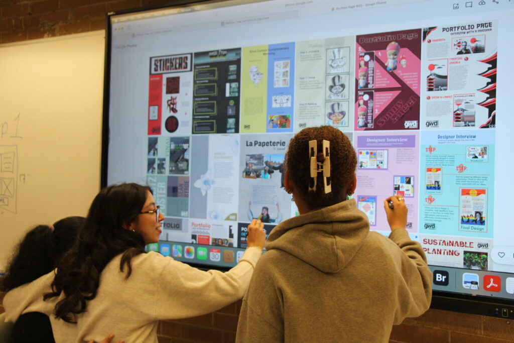 Three students looking at projects on the smart board in class P220 made by students from previous semesters from the Graphic & Web Design program.